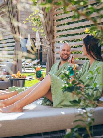 Eine Frau und ein Mann liegen draussen im Bademantel auf den Daybeds und geniessen einen Drink.