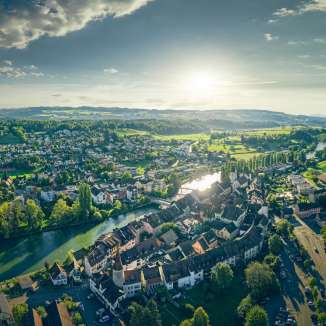 weite luftaufnahme von mellingen im aargau mit blick auf die reuss, die historische altstadt und die umliegenden felder und huegel im warmen licht.
