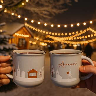 Zwei Hände halten weisse Glühweintassen mit Aarau-Motiv vor einem stimmungsvoll beleuchteten Weihnachtsmarkt mit Lichterketten.