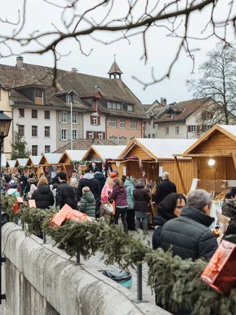 Besucherinnen und Besucher schlendern zwischen festlich geschmückten Holzhütten über die historische Rheinbrücke an der Altstadtweihnacht in Laufenburg.