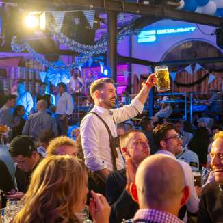 ein gast in tracht hebt seinen bierkrug ueber einer gut besuchten festgesellschaft mit blau-weisser dekoration und farbiger beleuchtung.