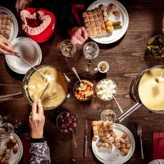 Mehrere Personen geniessen gemeinsam Käsefondue mit Brot und Beilagen an einem gedeckten Holztisch im Tägi Wettingen