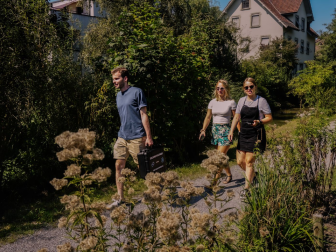 Drei Personen mit einem Rätselkoffer spazieren auf einem Weg entlang von Büschen und Wildblumen, im Hintergrund ein Wohnhaus.