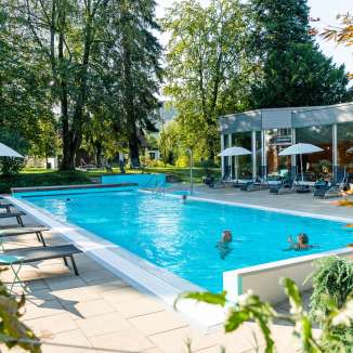 Aussenpool im Eden Spa mit türkisfarbenem Wasser, umgeben von Bäumen und Liegestühlen, im Hintergrund die Glasfassade des Spa-Gebäudes; mehrere Personen baden im Pool.