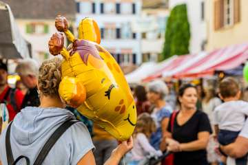Frau von hinten auf einem belebten Strassenmarkt, die einen gelben Giraffen-Folienballon in der Hand hält.