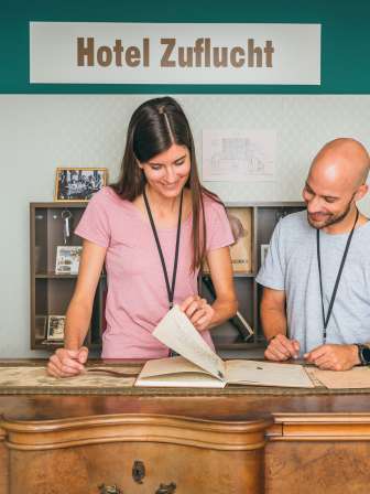 Eine Frau und eine Mann stehen im Historischen Museum hinter einer alten Theke und blättern im Reservationsbuch vom historischen Hotel Zuflucht.