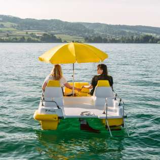 Zwei Personen fahren auf einem gelb-weissen Pedalboot mit Sonnenschirm ueber den Hallwilersee im Aargau. Das ruhige Wasser, die gruene Uferlandschaft und die sanfte Bergkulisse betonen die sommerliche, erholsame Stimmung.