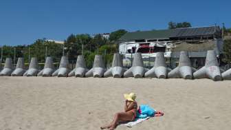 Chornomorsk – 20.06.2025: Eine Frau ruht am Strand von Chornomorsk, hinter ihr Beton-Panzerabwehrhindernisse entlang der Küste © Nataliia Mykhailenko / ukrainianphotodiary.org