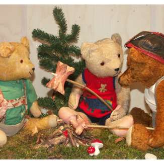 Drei Teddybaeren sitzen an einem kleinen Lagerfeuer und roesten gemeinsam Stockbrot.