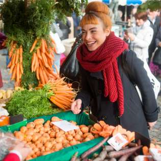 Lachende Frau mit rotem Schal kauft frische Karotten an einem bunt dekorierten Marktstand auf dem Aarauer Rüeblimärt.