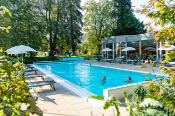 Aussenpool im Eden Spa mit türkisfarbenem Wasser, umgeben von Bäumen und Liegestühlen, im Hintergrund die Glasfassade des Spa-Gebäudes; mehrere Personen baden im Pool.