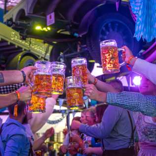 mehrere gaeste stossen mit gefuellten bierkruegen in einer lebhaften, farbenfroh beleuchteten oktoberfest halle gemeinsam an.