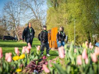 Drei Foxtrail-Teilnehmende spazieren durch den Park bei der Aare in Aarau. Frühlingsblumen und grüne Bäume bilden den farbenfrohen Rahmen des Stadtspiels.