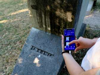 Hand hält Smartphone mit Rätsel-App vor einem Steindenkmal mit Inschrift im Grünen.