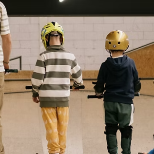 Familie im Rolling Rock Skate- & Sportcenter, 3 Kinder stehen mit Scootern vor einer Rampe und warten auf die Abfahrt.
