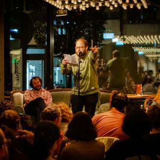 Poetry-Slammer trägt mit Mikrofon und Textblatt vor einem sitzenden Publikum im tibits vor.