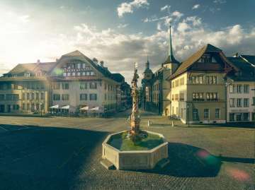 Das Bild zeigt den Niklaus-Thut-Brunnen im Zentrum der historischen Altstadt von Zofingen in der Schweiz. Der kunstvoll gestaltete Brunnen steht auf einem weitlaeufigen Marktplatz und wird von gut erhaltenen, traditionellen Altstadthaeusern mit Giebeldaechern und Fassaden aus verschiedenen Epochen umrahmt. Im Hintergrund fuehren schmale Gassen durch die Altstadt, waehrend das warme Morgen- oder Abendlicht die Architektur und den Platz in eine ruhige, atmosphaerische Stimmung taucht. Der wolkige Himmel verleiht der Szene Tiefe und unterstreicht den historischen Charakter des Ortes.