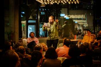 Poetry-Slammer trägt mit Mikrofon und Textblatt vor einem sitzenden Publikum im tibits vor.