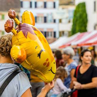 Frau von hinten auf einem belebten Strassenmarkt, die einen gelben Giraffen-Folienballon in der Hand hält.