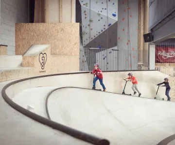 Drei Kinder fahren mit Scootern durch einen Indoor-Bowl im Rolling Rock Aarau, im Hintergrund befindet sich eine Kletterwand.