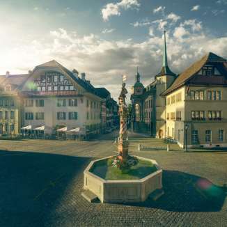 Das Bild zeigt den Niklaus-Thut-Brunnen im Zentrum der historischen Altstadt von Zofingen in der Schweiz. Der kunstvoll gestaltete Brunnen steht auf einem weitlaeufigen Marktplatz und wird von gut erhaltenen, traditionellen Altstadthaeusern mit Giebeldaechern und Fassaden aus verschiedenen Epochen umrahmt. Im Hintergrund fuehren schmale Gassen durch die Altstadt, waehrend das warme Morgen- oder Abendlicht die Architektur und den Platz in eine ruhige, atmosphaerische Stimmung taucht. Der wolkige Himmel verleiht der Szene Tiefe und unterstreicht den historischen Charakter des Ortes.