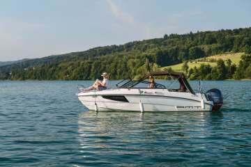 Zwei Personen geniessen eine entspannte Fahrt auf einem modernen Motorboot auf dem Hallwilersee im Aargau. Das klare Wasser und die bewaldeten Huegel im Hintergrund schaffen eine ruhige Sommerstimmung.